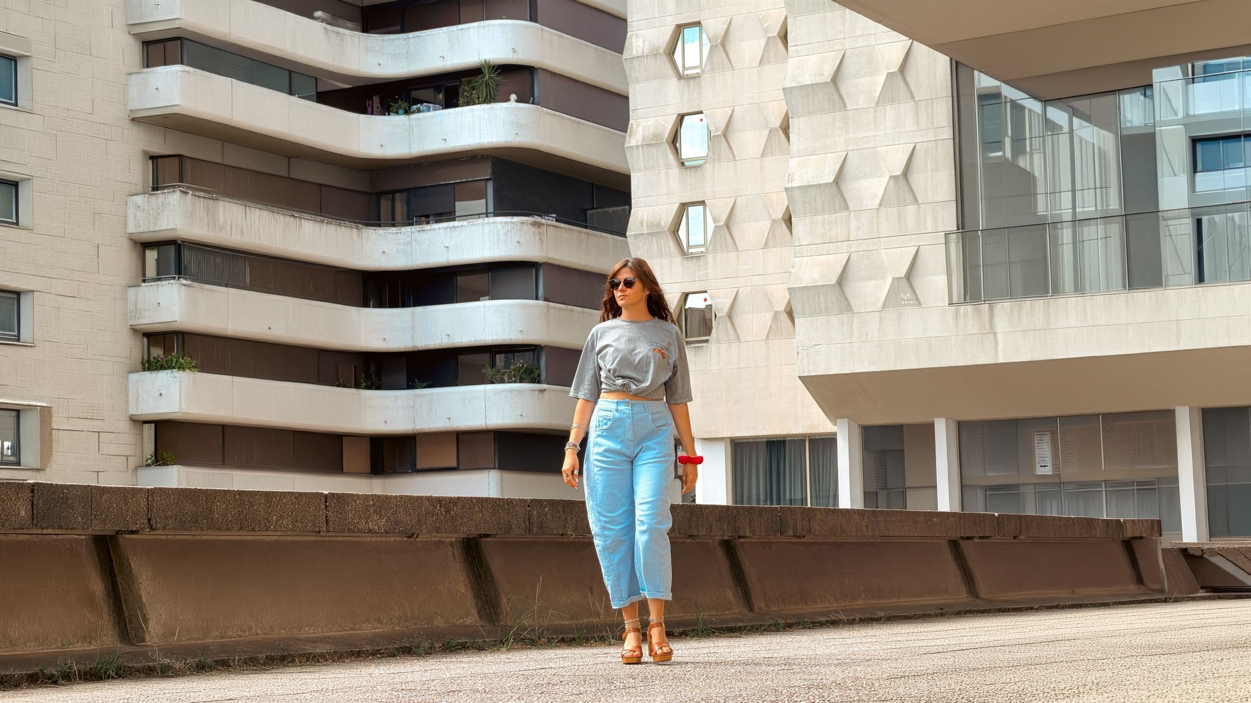 Femme en t-shirt gris noué et jean clair devant une façade d'immeuble moderne