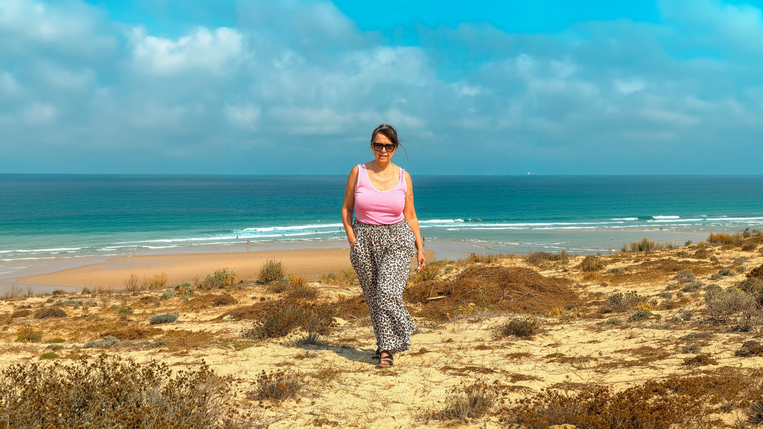 Femme en débardeur rose et pantalon léopard marchant sur une dune face à l'océan