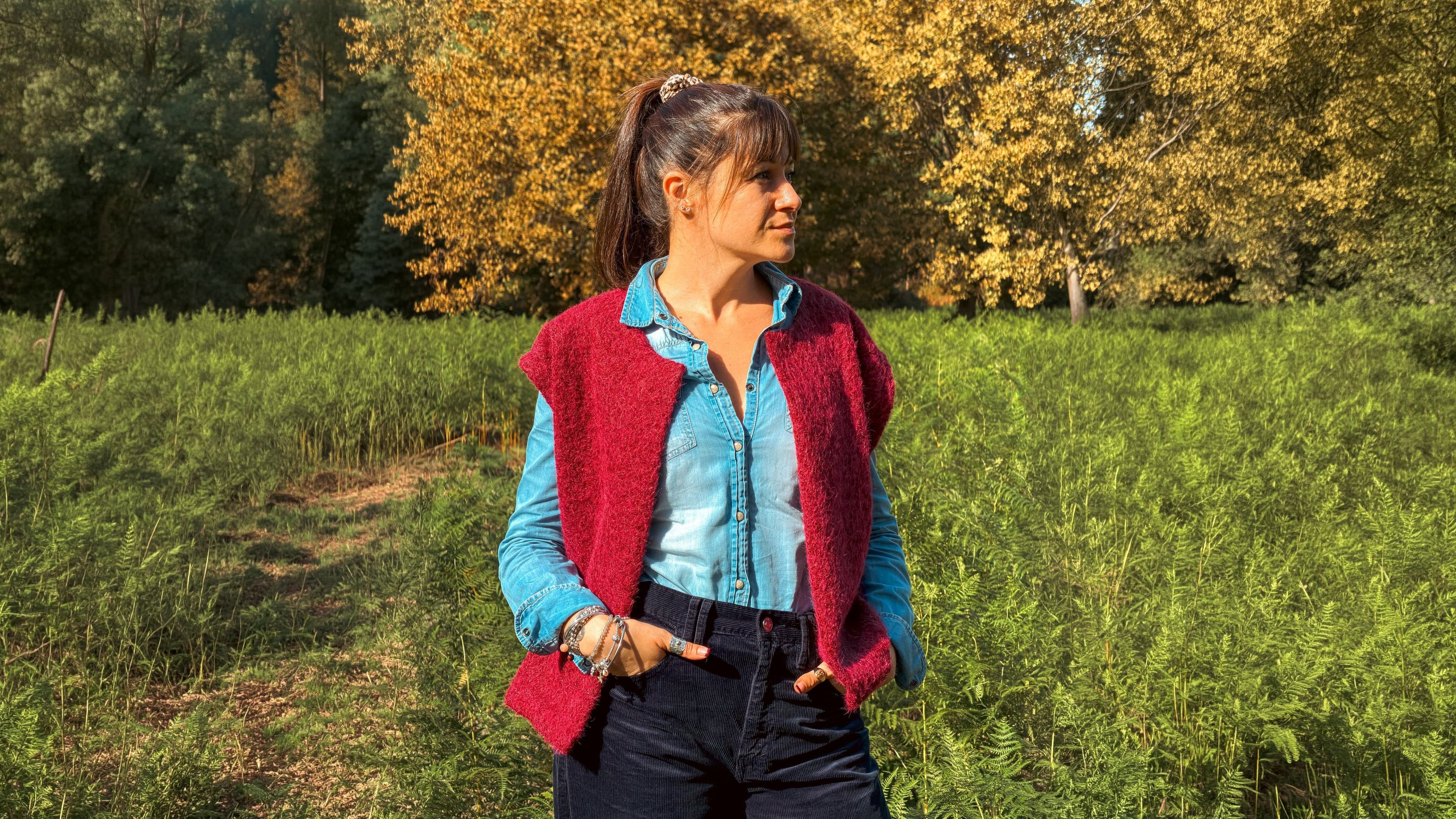 Femme en gilet sans manches en maille rouge et chemise en jean, mains dans les poches, au milieu des fougères sous les arbres d'automne