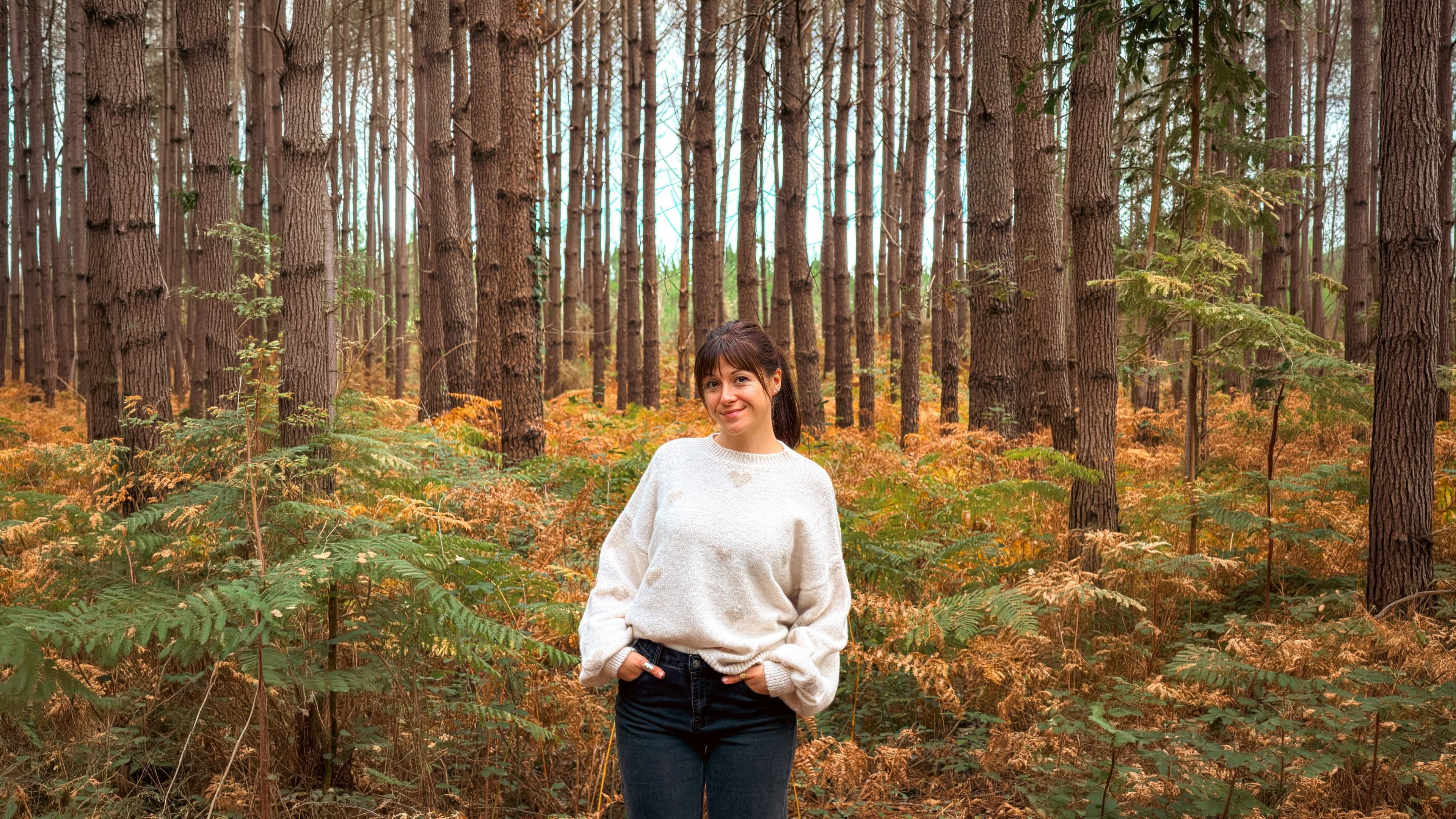 Femme souriante en pull en maille blanc cassé brodé de cœurs et jean brut, debout dans une forêt de pins parmi les fougères dorées